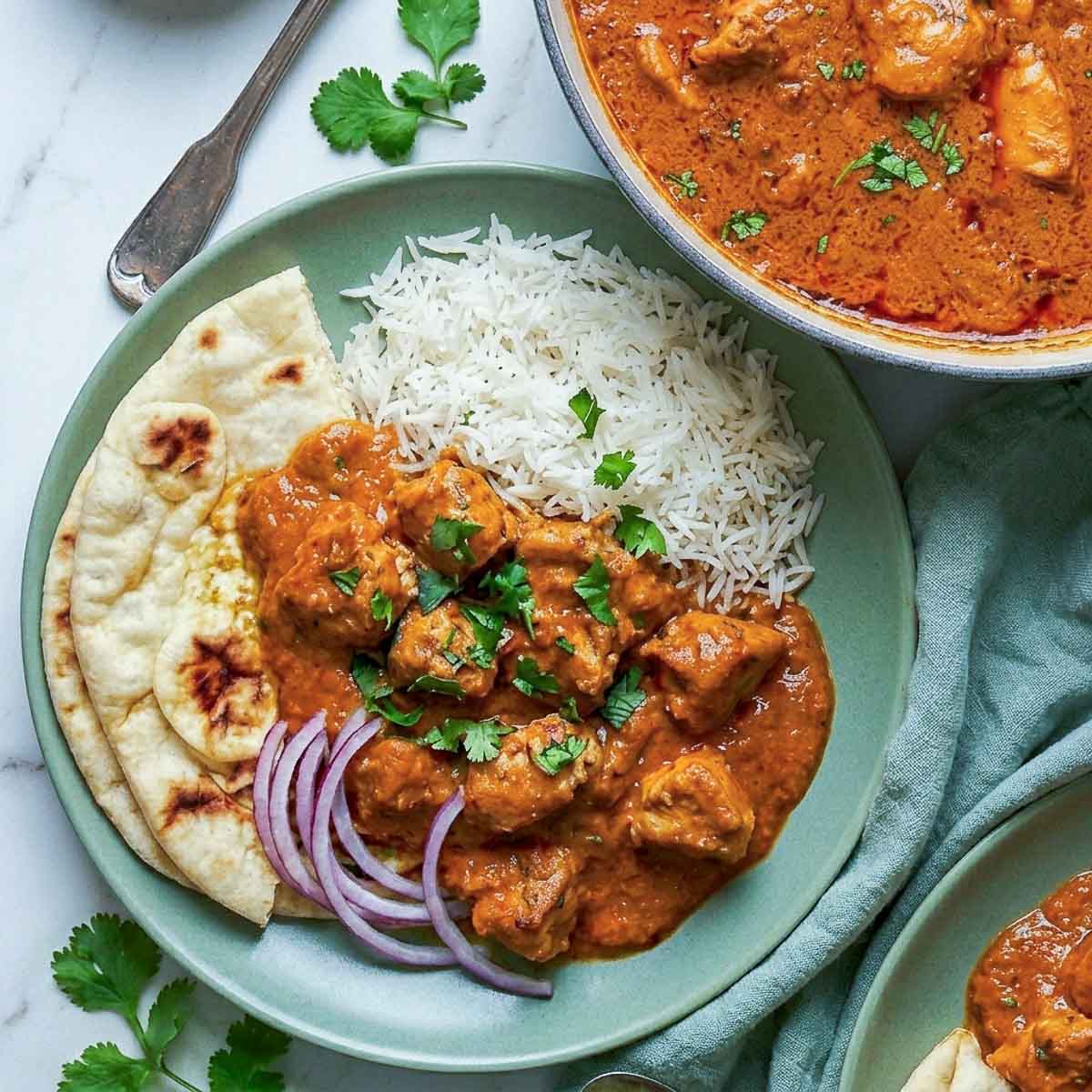 Overhead view of a plated serving of chicken masala with basmati rice, naan, sliced red onions, and fresh cilantro, with the Dutch oven visible in the background.