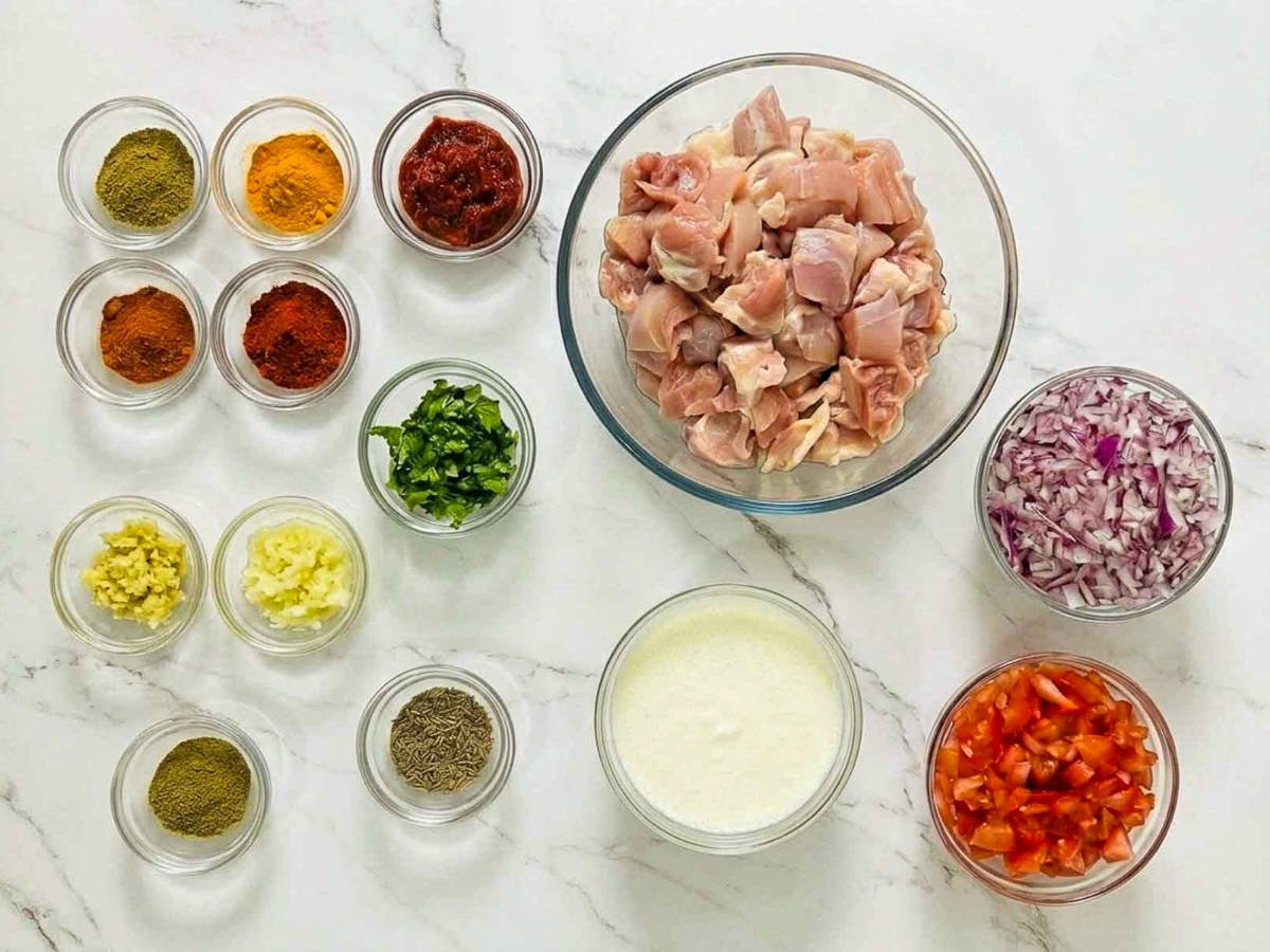Top-down view of chicken masala ingredients including raw chicken thighs, chopped tomatoes and onions, yogurt, tomato paste, ginger, garlic, cilantro, cumin seeds, and Indian spices arranged on white marble.