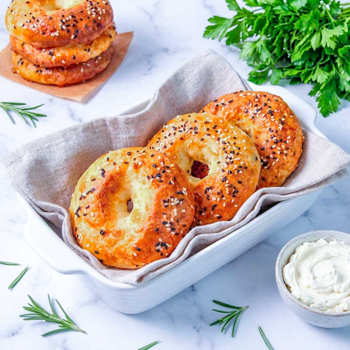 Close-up of four cottage cheese bagels topped with everything bagel seasoning in a linen-lined white baking dish, with a bowl of cream cheese alongside.