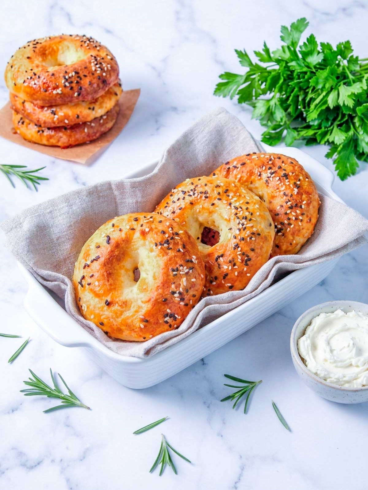 Four cottage cheese bagels topped with everything bagel seasoning in a linen-lined white baking dish, with a bowl of cream cheese alongside.