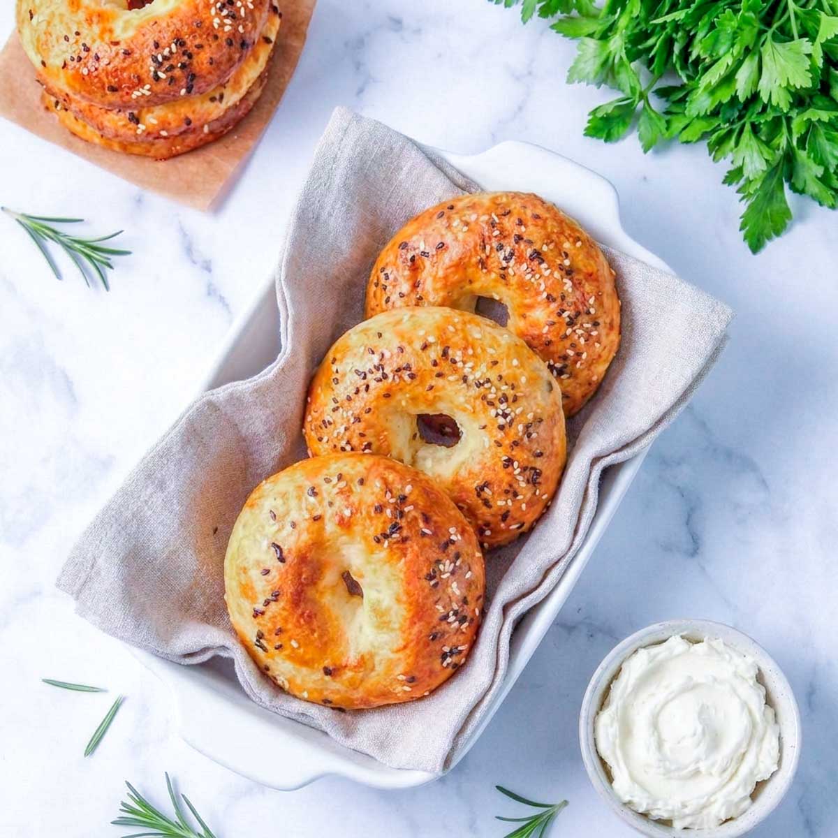 Overhead view of three cottage cheese bagels in a linen-lined dish with a bowl of cream cheese and fresh herbs.