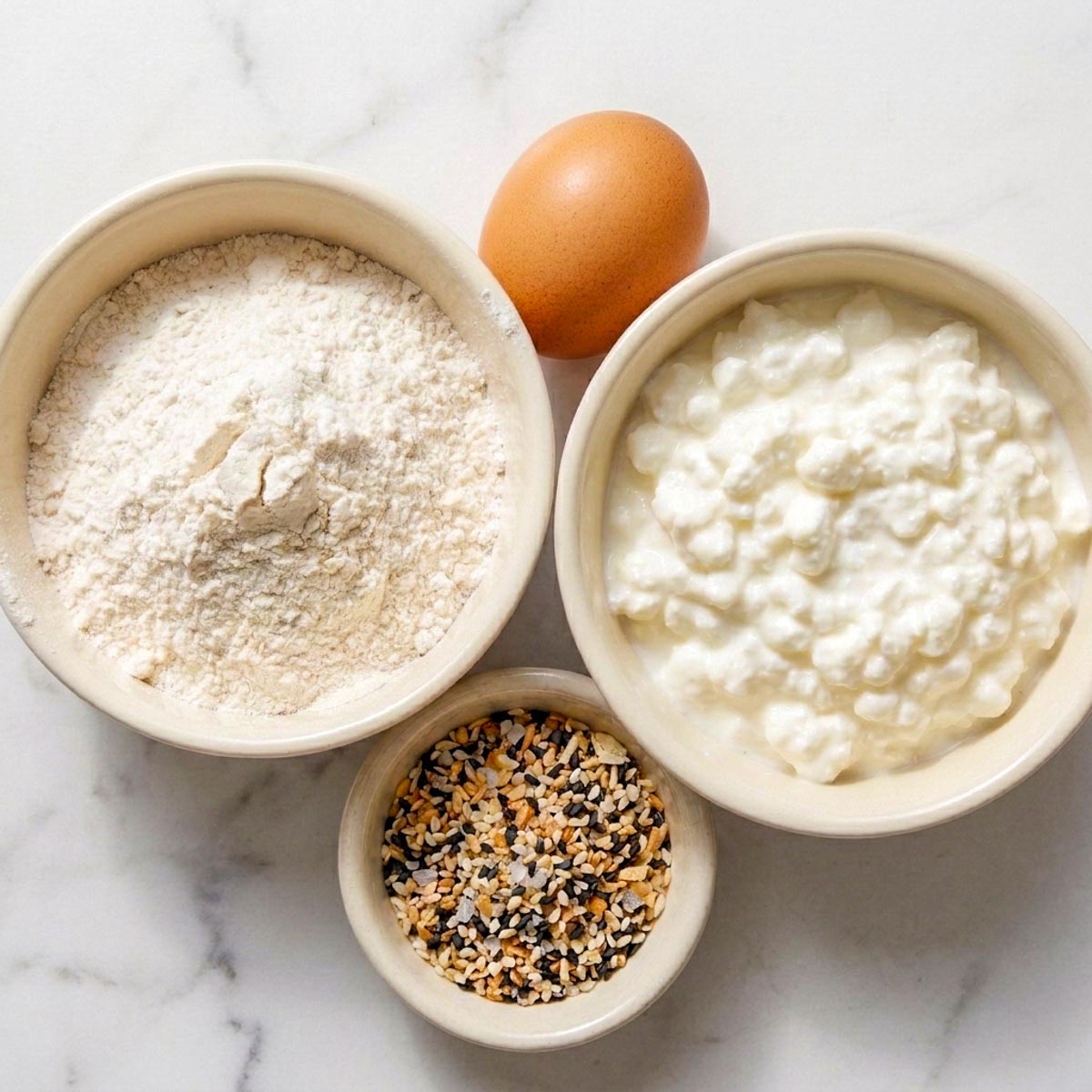 Self-rising flour, cottage cheese, a brown egg, and everything bagel seasoning in ceramic bowls on a marble surface.