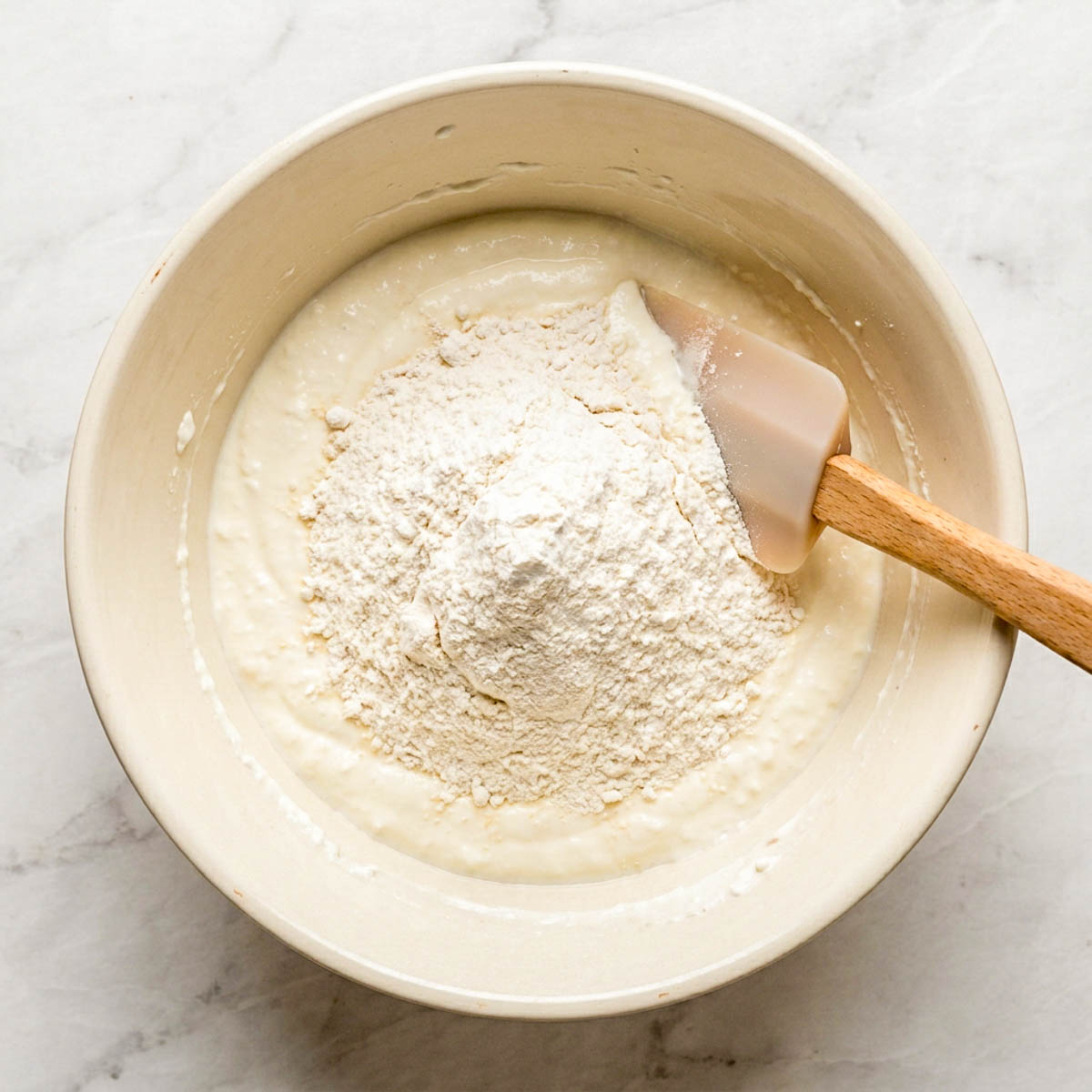 Flour added to blended cottage cheese in a cream ceramic bowl with a spatula.