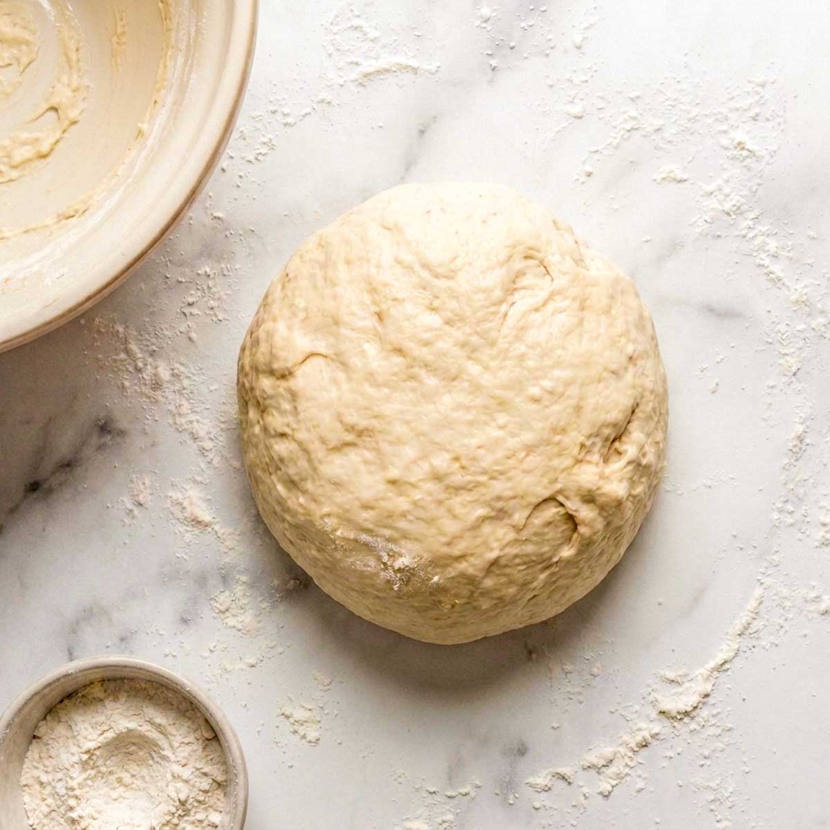 Smooth ball of cottage cheese bagel dough on a floured marble surface next to the mixing bowl.