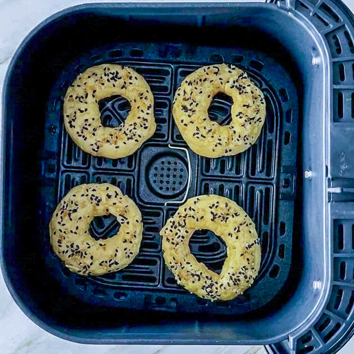 Four raw cottage cheese bagels topped with seeds arranged in a black air fryer basket.