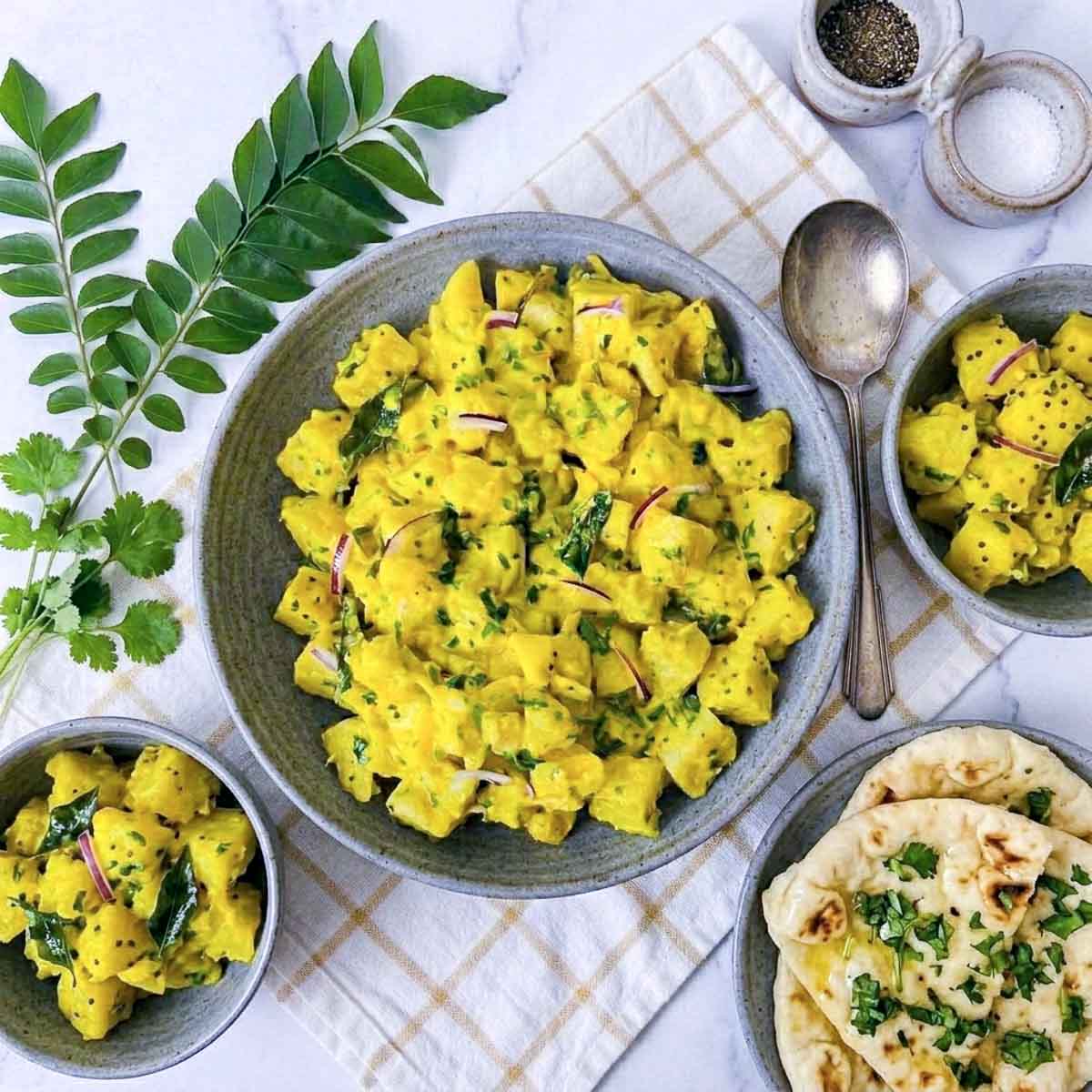 Curry potato salad in a gray bowl with curry leaves, red onion, and warm naan alongside.