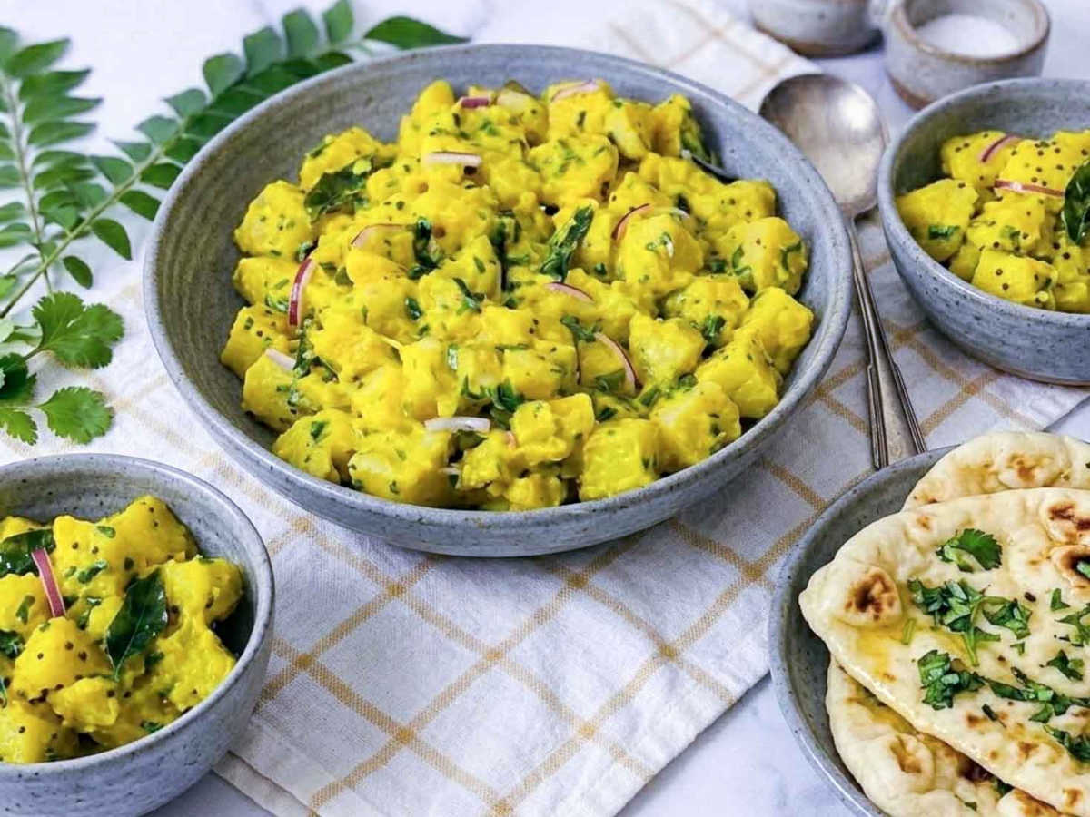 Curry potato salad in a large gray ceramic serving bowl, garnished with cilantro, curry leaves, red onion, and black mustard seeds, with naan and additional bowls of potato salad on a checked linen napkin.