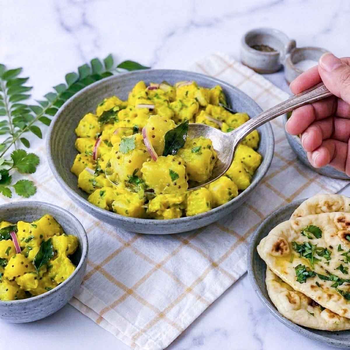 Curried potato salad served in a gray ceramic bowl with a spoonful lifted, garnished with cilantro, red onion, curry leaves, and mustard seeds.