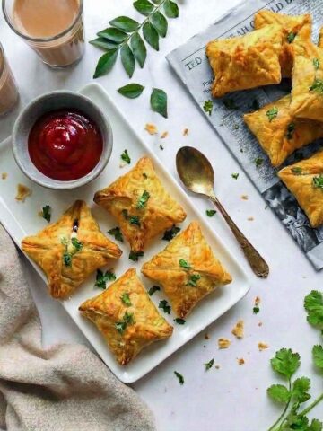 An overhead photo of Indian egg puffs arranged on a white platter with ketchup, two glasses of chai, fresh curry leaves, and cilantro.