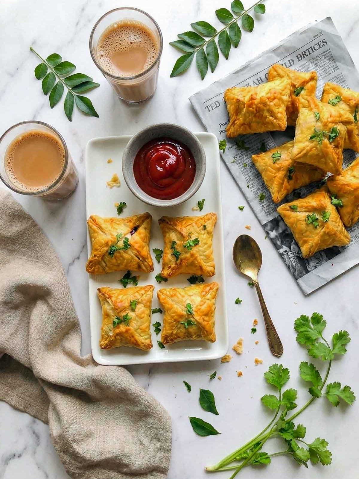 An overhead photo of homemade egg puffs arranged on a white platter with ketchup, two glasses of chai, fresh curry leaves, cilantro, and a spoon.