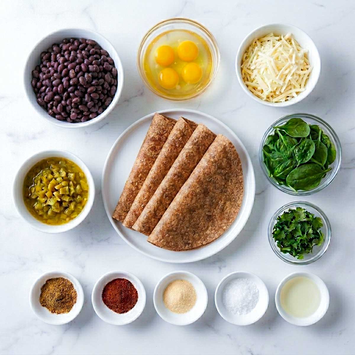 Ingredients for freezer breakfast quesadillas in white bowls on marble, including black beans, eggs, shredded cheese, spinach, green chiles, and spices.