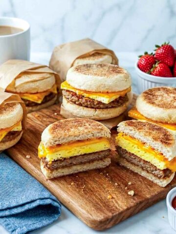Freezer breakfast sandwiches on a wooden board with two cut in half showing egg, turkey sausage, and melted cheddar layers.
