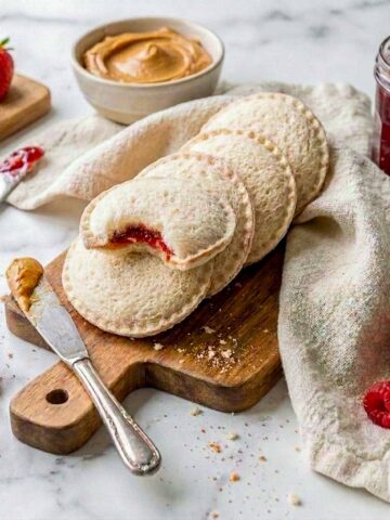 Homemade uncrustables on a wooden board with peanut butter and jam, one cut open to show the filling.