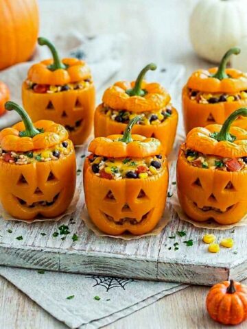 Six orange jack-o'-lantern stuffed peppers filled with rice, black beans, corn, tomatoes, and melted cheese on a rustic wooden board.