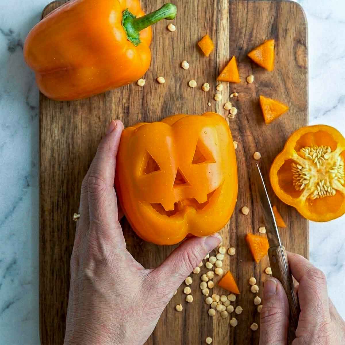 Hands carving a jack-o'-lantern face into an orange bell pepper on a wooden cutting board, with pepper seeds and cut pieces nearby.
