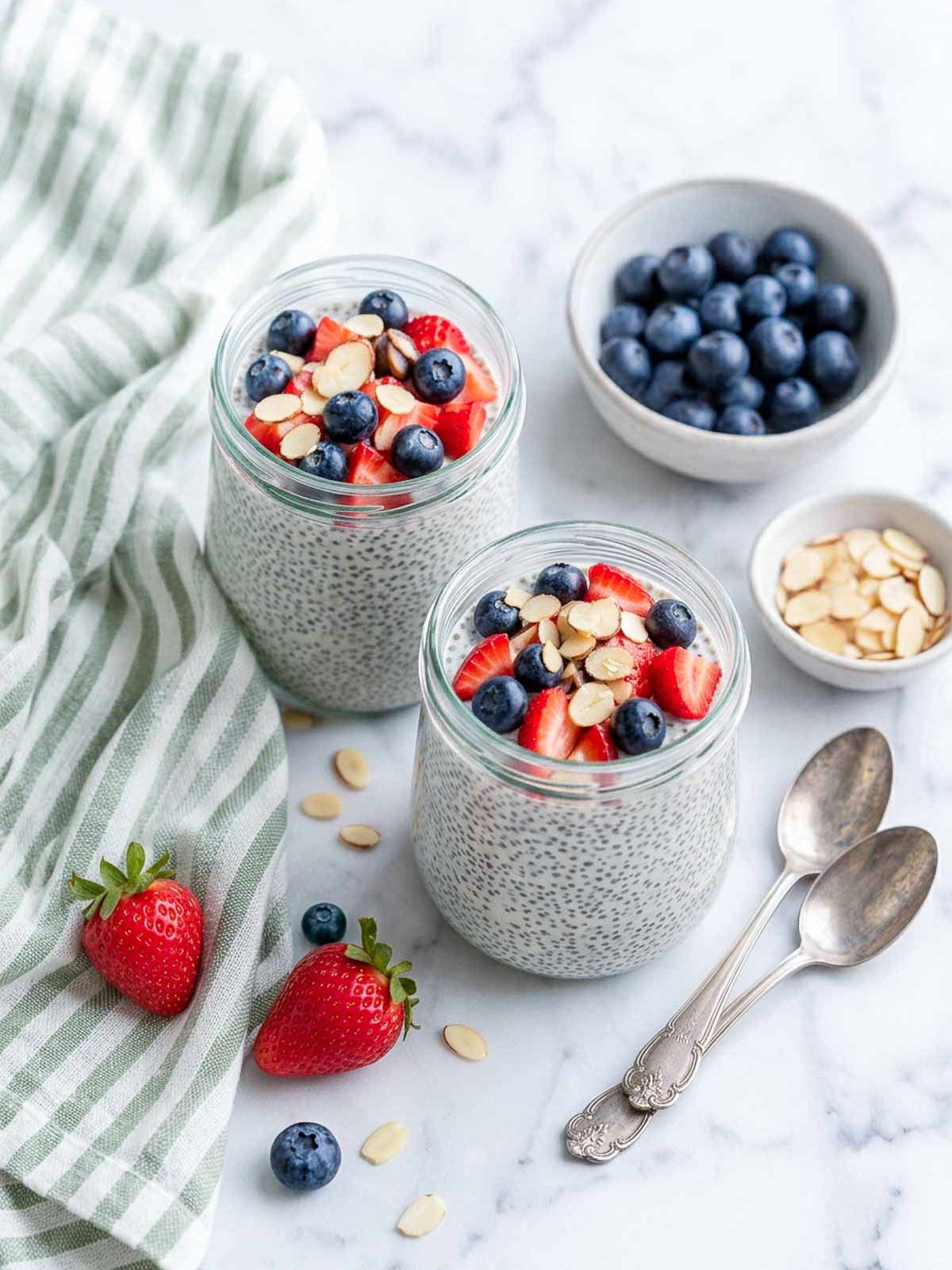 Kefir chia pudding in two glass jars with fresh strawberries, blueberries, and sliced almonds beside a green striped napkin.