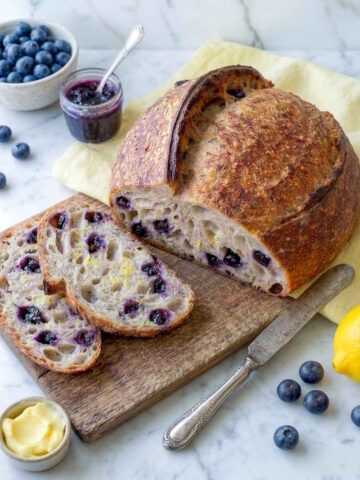 Lemon blueberry sourdough bread boule with two slices cut, blueberries, jam, and a lemon alongside.
