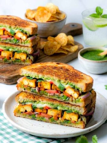 Paneer sandwich cut in half and stacked on a plate with green chutney, lacha onion, and chips.