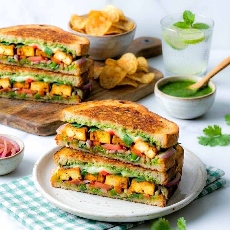 Paneer sandwich cut in half and stacked on a plate with green chutney, lacha onion, and chips.