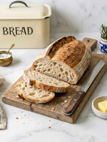 Sliced same day sourdough bâtard on a wooden board with a serrated knife, butter dish and cream enamel bread box.