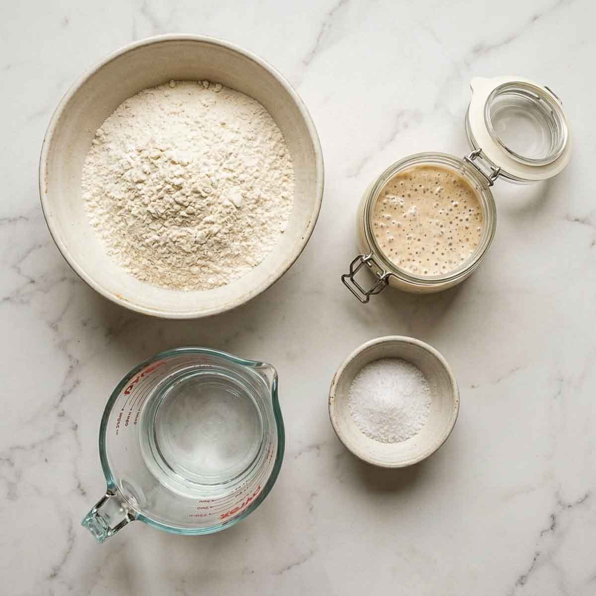 Bread flour, bubbling sourdough starter, water and fine sea salt in ceramic bowls on white marble.