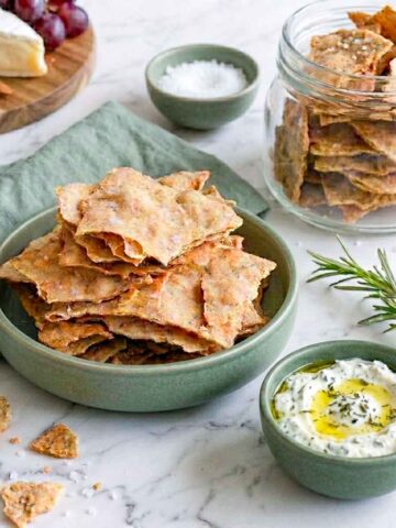 Bowl of thin golden sourdough discard crackers with a storage jar, flaky sea salt, and herb dip.
