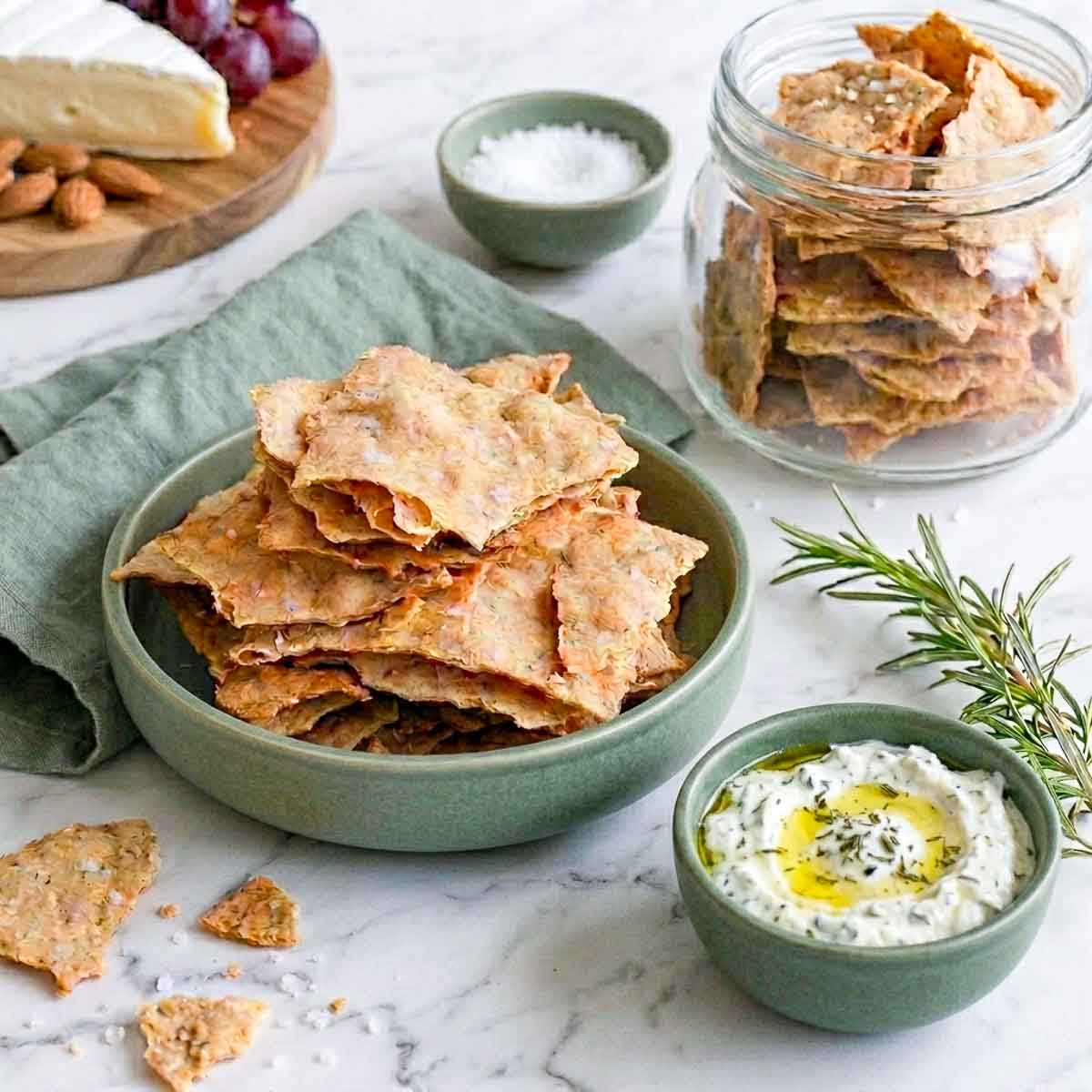 Bowl of thin golden sourdough discard crackers with a storage jar, flaky sea salt, and herb dip.