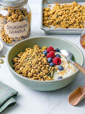 Sourdough granola served in a sage green bowl with yogurt, fresh berries, and a baking tray of granola in the background.