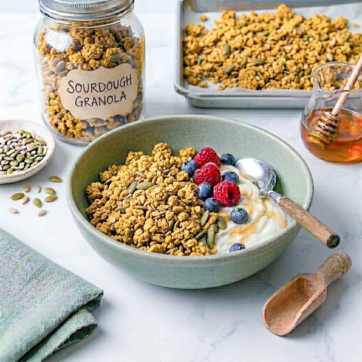 Sourdough granola served in a sage green bowl with yogurt, fresh berries, and a baking tray of granola in the background.