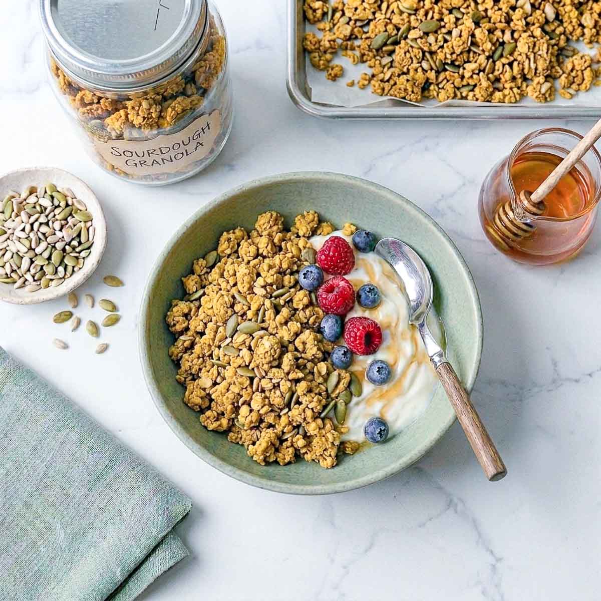 Overhead view of sourdough granola served with yogurt, raspberries and blueberries in a green bowl, with a jar of granola and a dish of seeds alongside.