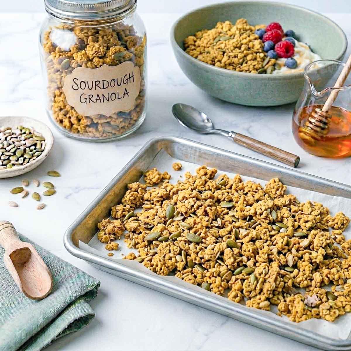 Sourdough granola cooling on a steel baking tray lined with parchment, with a jar of granola and honey alongside.