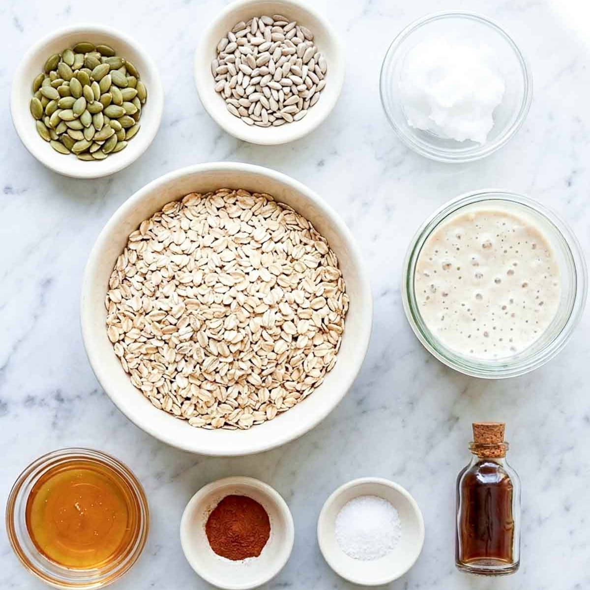 Sourdough granola ingredients laid out on marble: rolled oats, bubbly sourdough starter, pumpkin and sunflower seeds, honey, coconut oil, cinnamon, salt and vanilla.