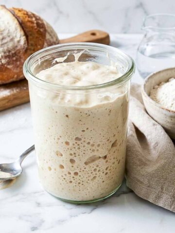 Active sourdough starter in a glass jar beside a bowl of bread flour and a finished loaf.