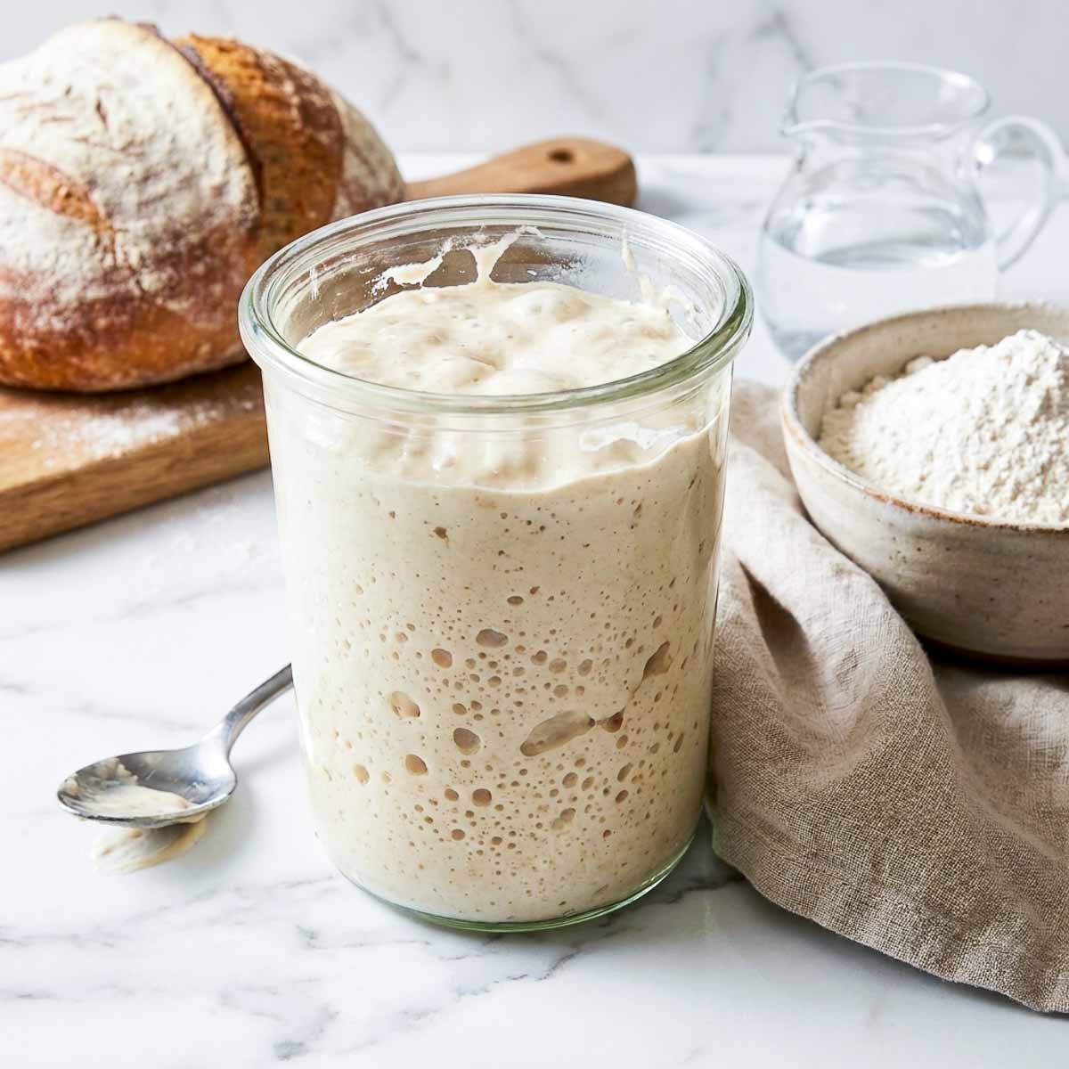 Active sourdough starter in a glass jar beside a bowl of bread flour and a finished loaf.