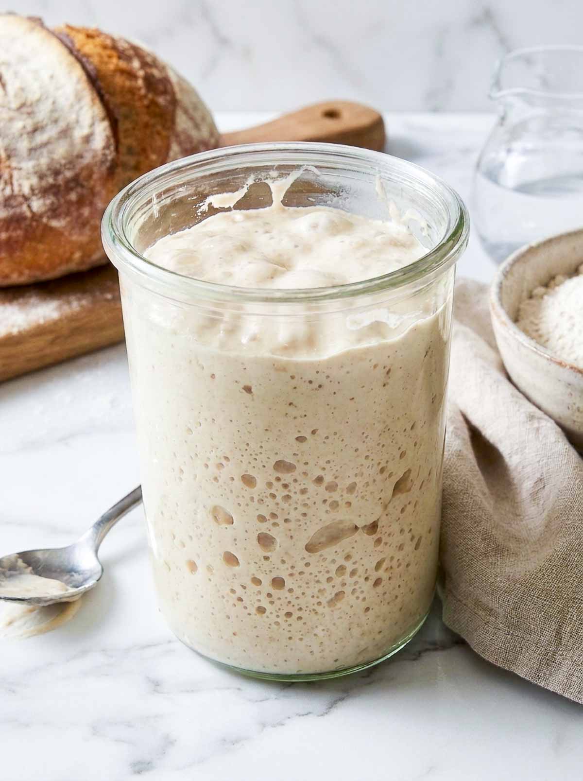 Close up of active sourdough starter in a glass jar beside a bowl of bread flour and a finished loaf.