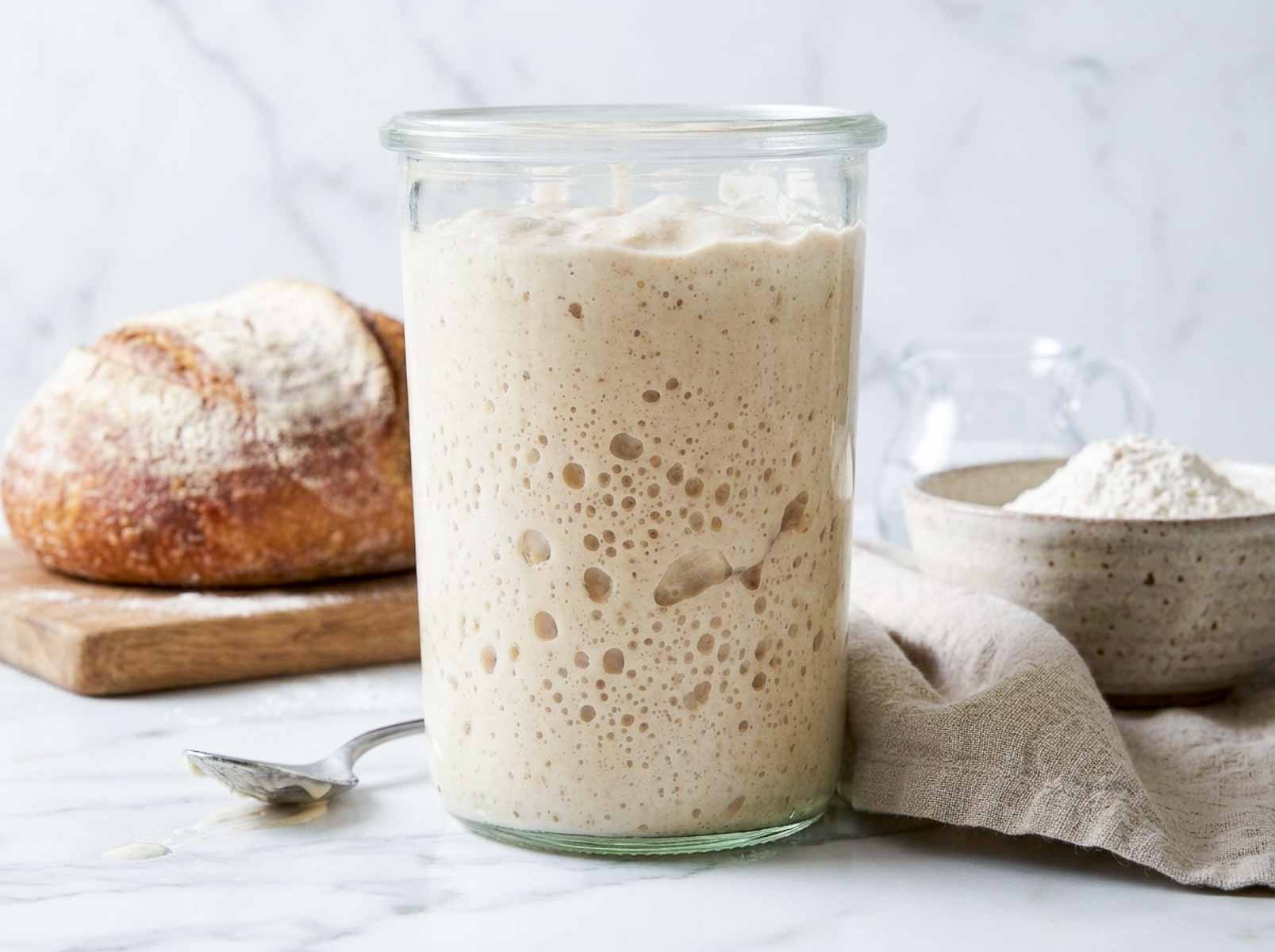 Sourdough starter in a glass jar on a marble counter with bread flour and a baked loaf.