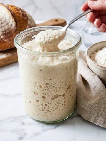 Spoon lifting bubbly sourdough starter from a glass jar while feeding it.