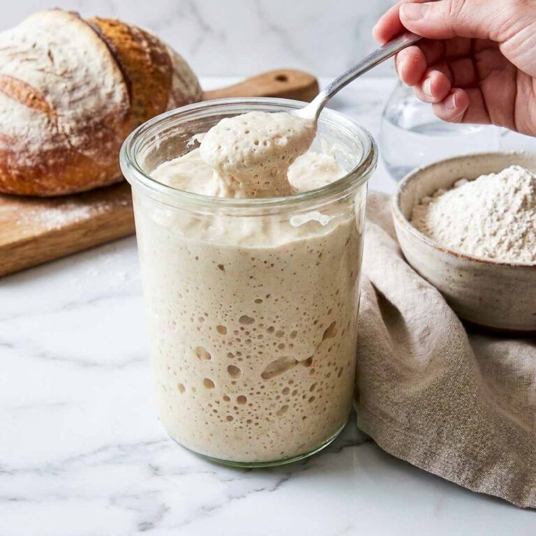 Spoon lifting bubbly sourdough starter from a glass jar while feeding it.