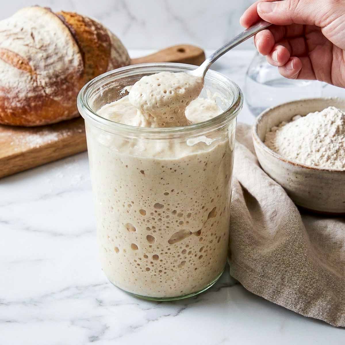 Spoon lifting bubbly sourdough starter from a glass jar while feeding it.