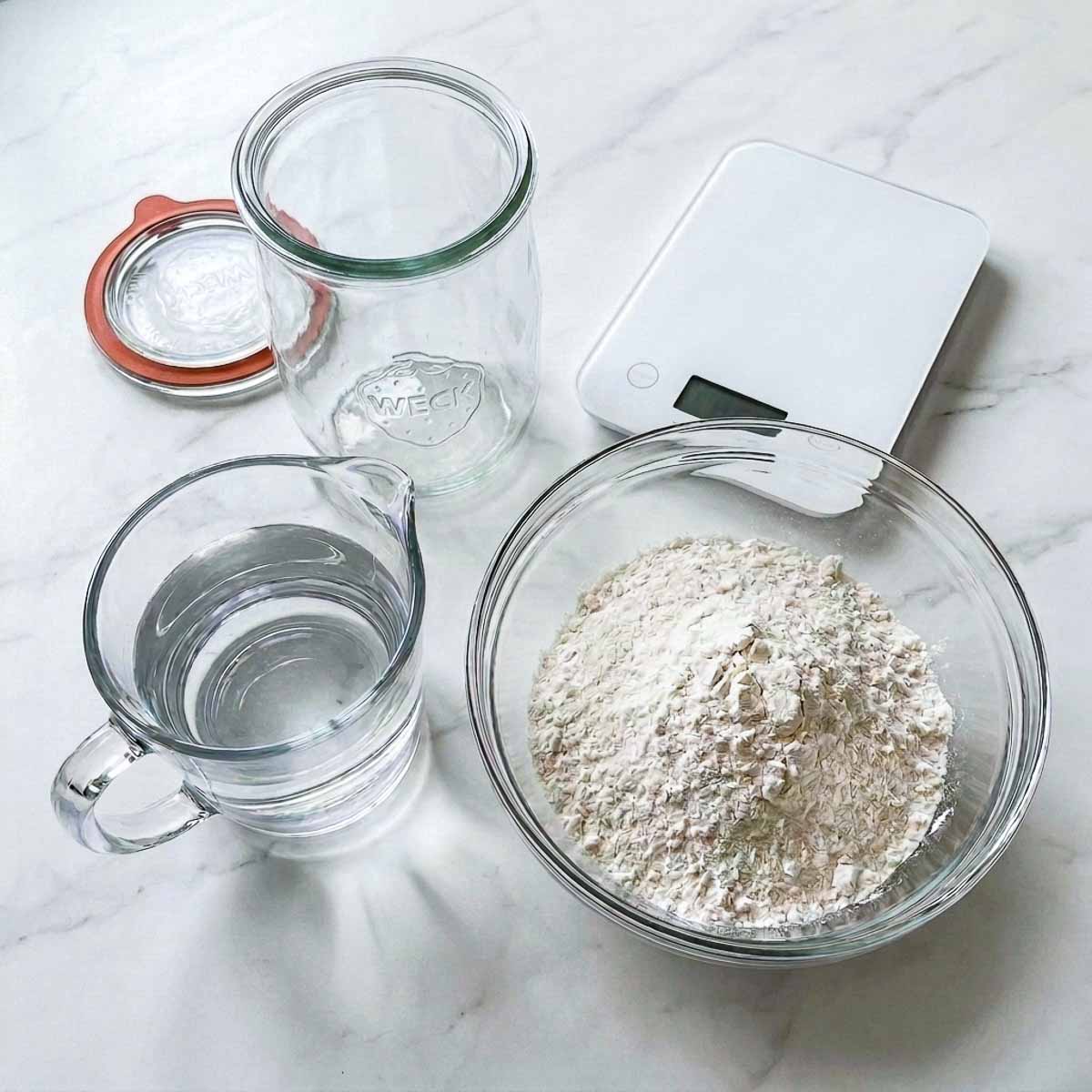 Bread flour, filtered water, an empty glass jar, and a kitchen scale for making sourdough starter.
