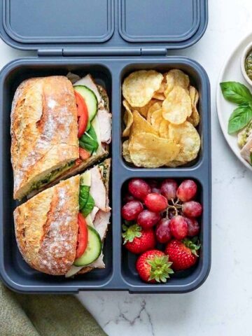 Turkey pesto baguette placed in a lunchbox along with fruits.
