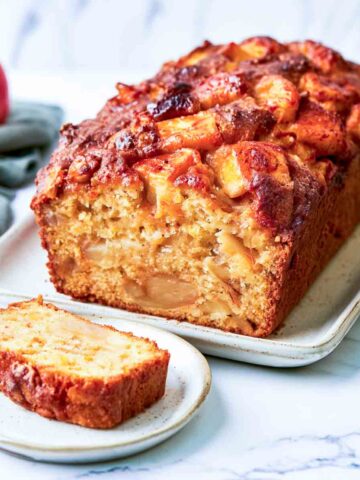 Close-up angled view of a kefir apple loaf cake showing its golden, apple-filled interior and caramelized apple pieces on top. A single slice rests on a small white plate in the foreground, with a sage green napkin, fresh apples, and stacked white plates visible around the loaf on a white marble surface.
