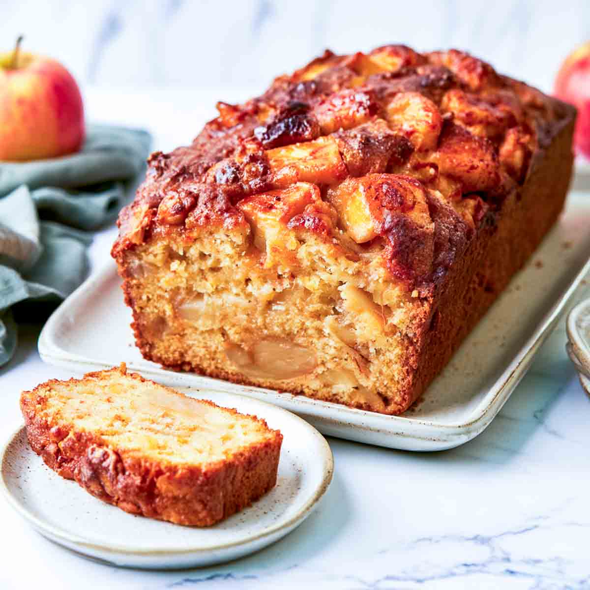 Close-up angled view of a kefir apple loaf cake showing its golden, apple-filled interior and caramelized apple pieces on top. A single slice rests on a small white plate in the foreground, with a sage green napkin, fresh apples, and stacked white plates visible around the loaf on a white marble surface.