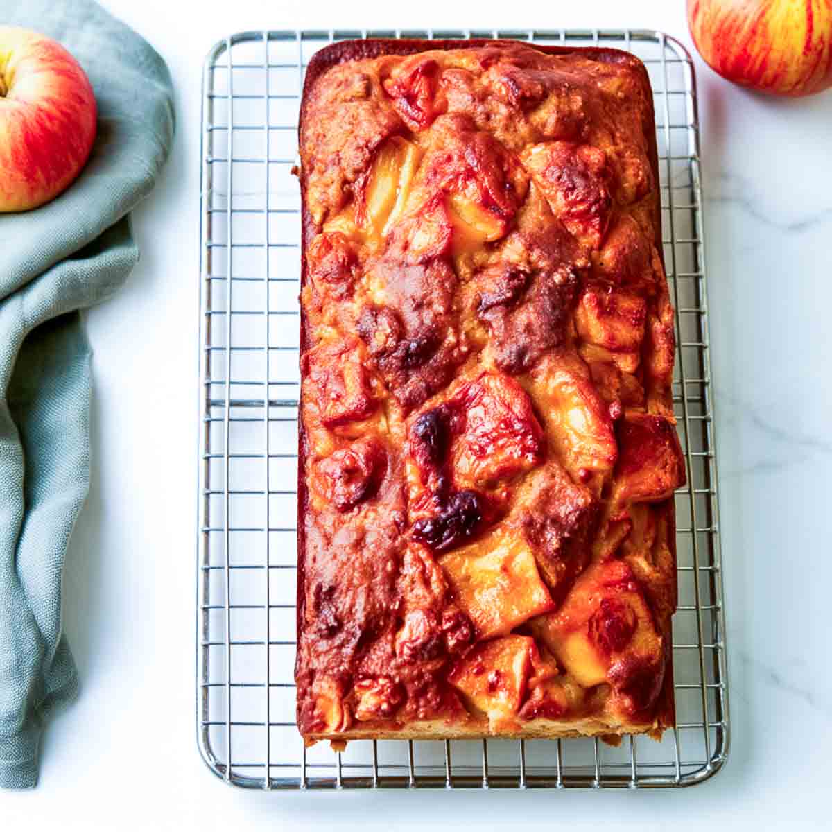 Whole kefir apple loaf cake cooling on a wire rack next to fresh apples and a sage green linen.