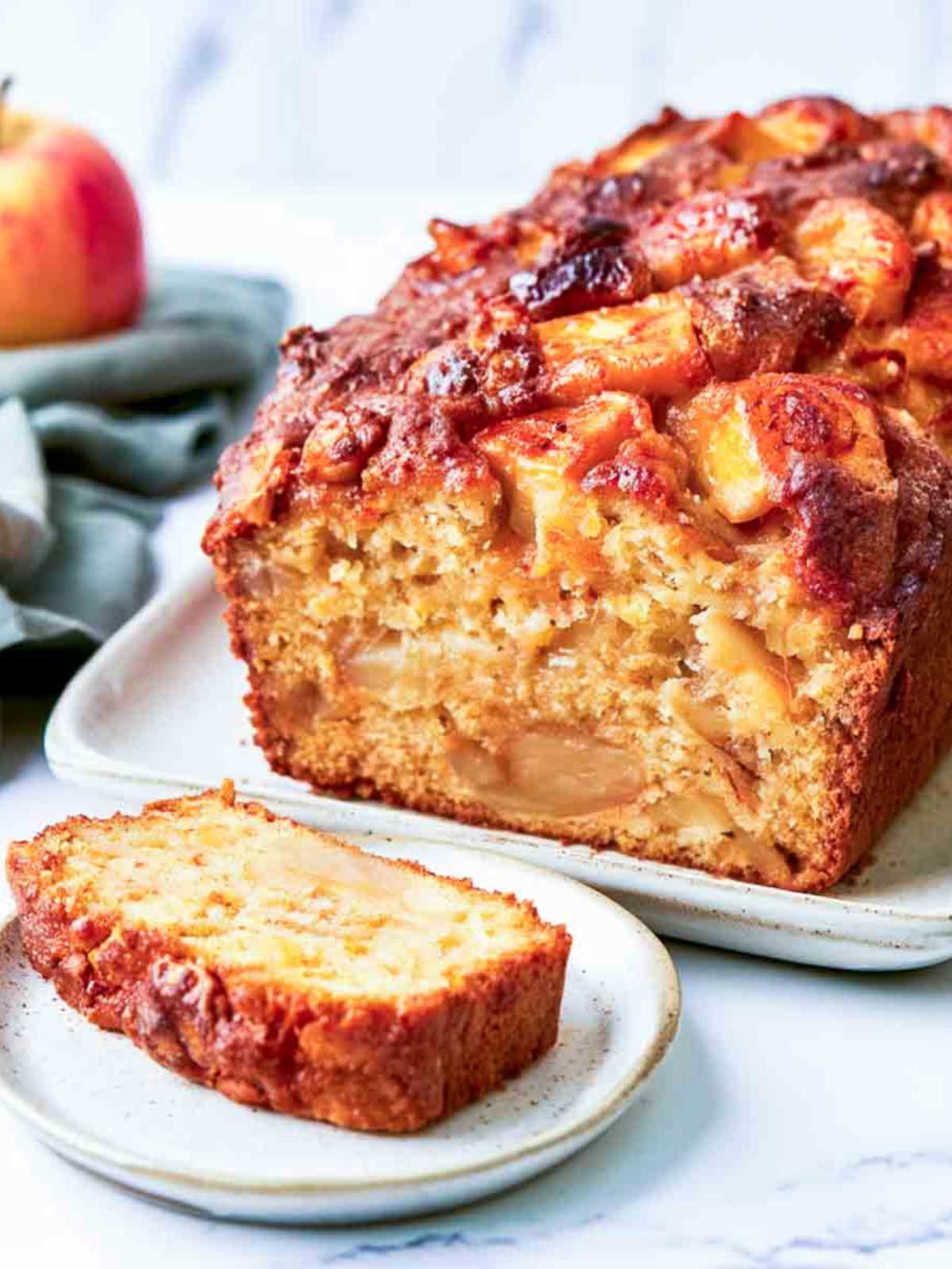 Close-up of a kefir apple loaf cake with a golden-brown crust and caramelized apple pieces on top, showing a moist apple-filled crumb. A single slice sits on a white plate in the foreground, with a sage green napkin and apple in the background on a white marble surface.