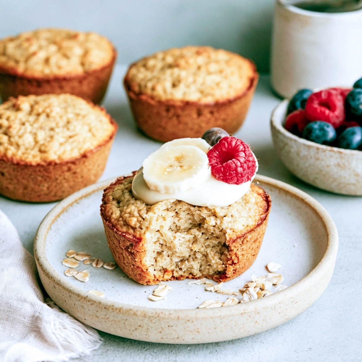 Close-up photograph of a golden-brown baked oatmeal cup topped with yogurt, banana slices, a raspberry, and a blueberry, served on a rustic ceramic plate. More oatmeal cups and a bowl of fresh berries are softly arranged in the background, with rolled oats scattered around the plate.