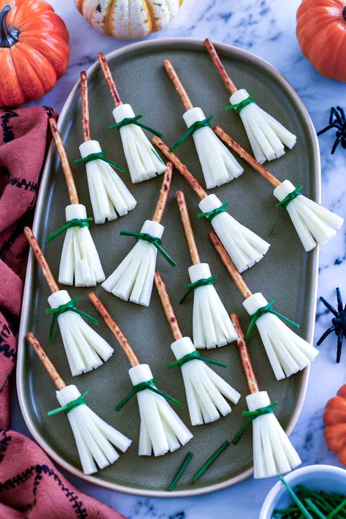 Cheese broomsticks made from string cheese and pretzel sticks tied with chives on a green platter.