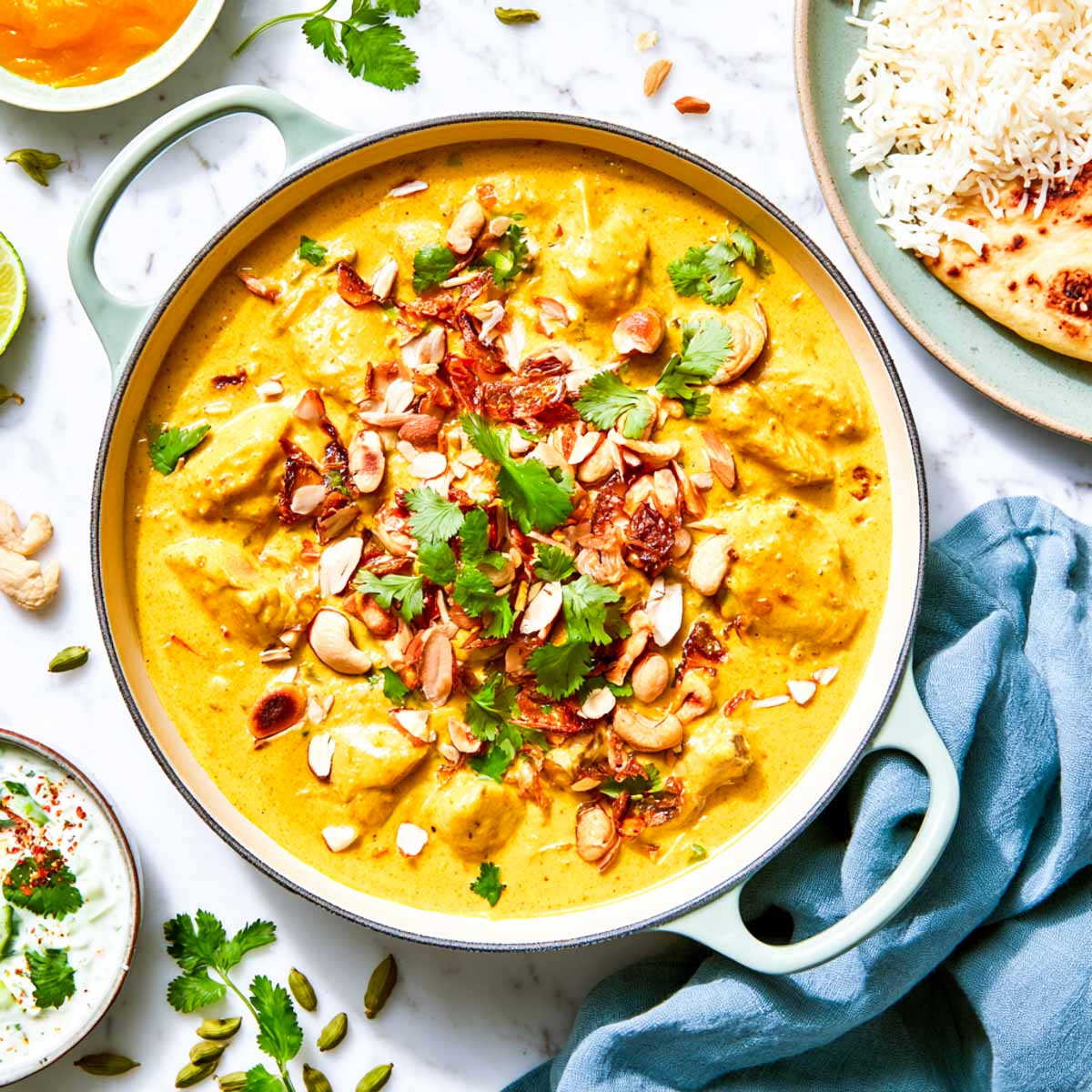 Close up of chicken korma in a pale green braiser topped with crispy fried onions, cashews, and cilantro.