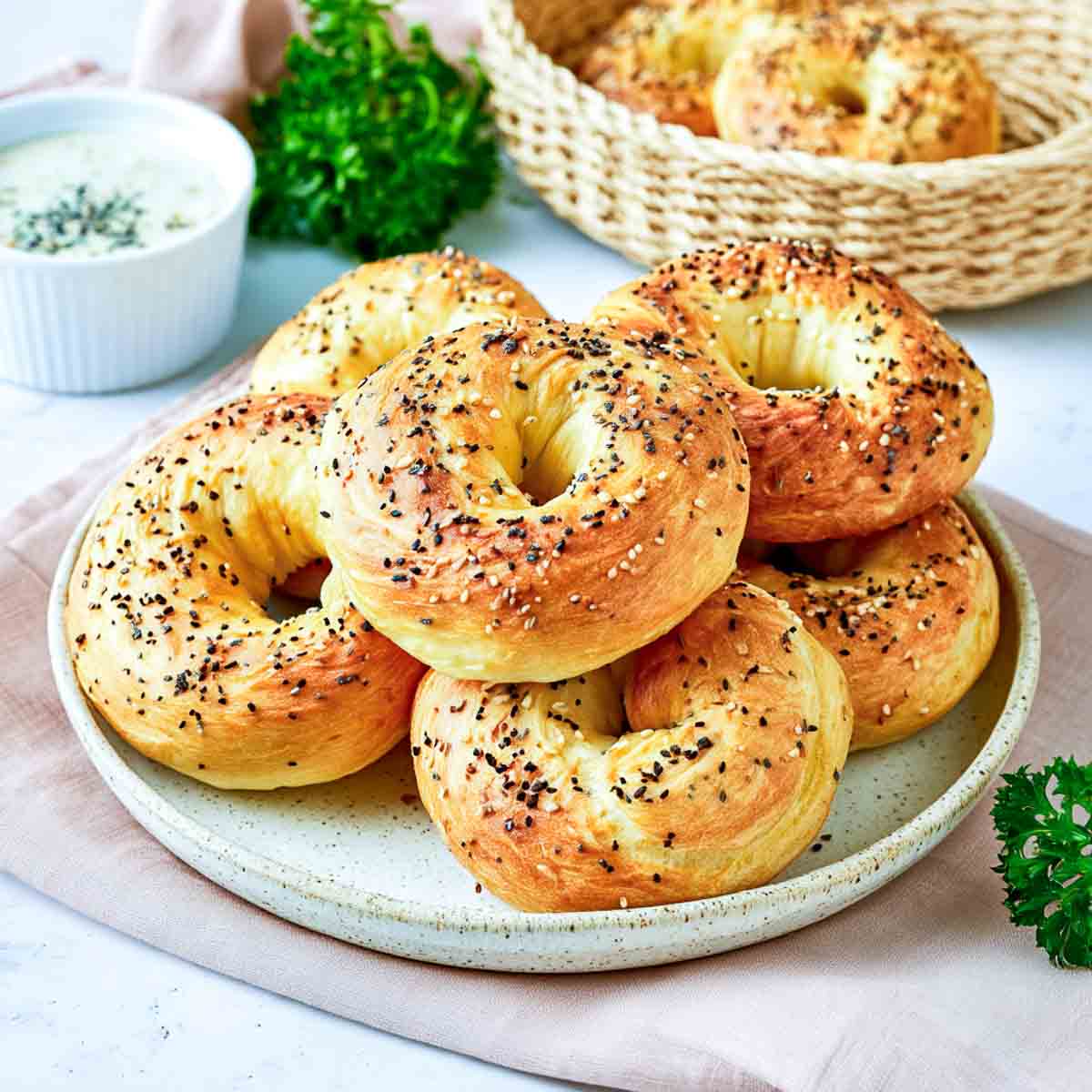 Homemade cottage cheese bagels topped with everything bagel seasoning, stacked on a ceramic plate with a basket of bagels and creamy dip in the background.