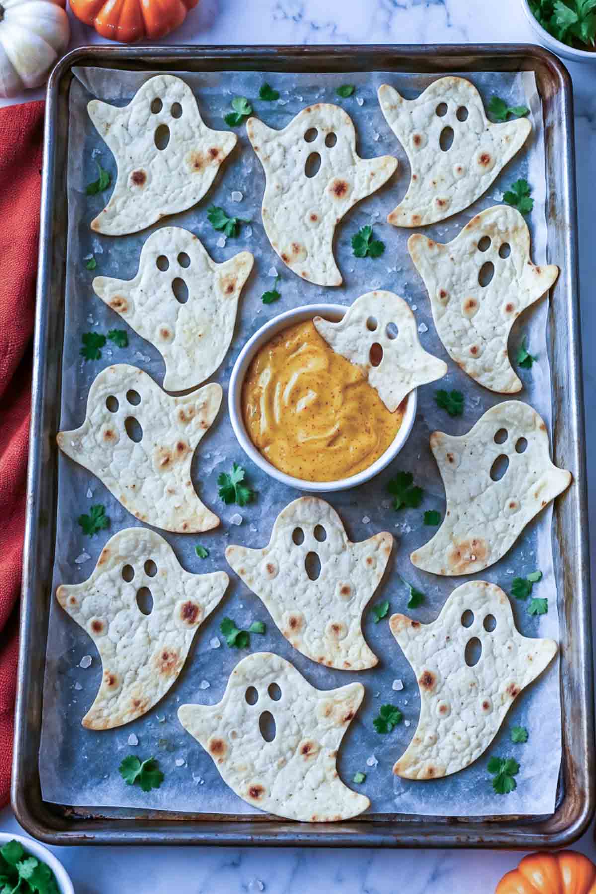 Ghost shaped tortilla chips on a sheet pan around a bowl of orange cheese dip.